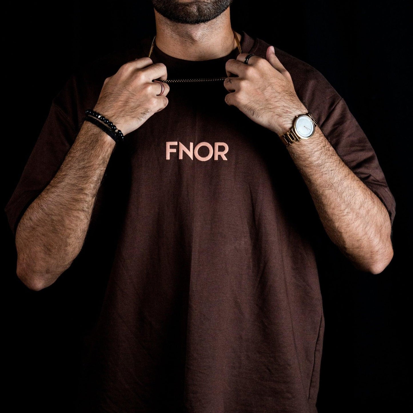 A man confidently pulling the collar of the TRIBAL GANGSTER BROWN T-SHIRT by FNOR, showcasing the FNOR logo in a bold puff print.