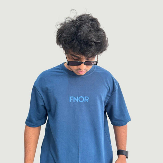 A man in an AIRFORCE blue FNOR t-shirt with the FNOR logo inscribed on the front, casually looking down while wearing sunglasses.