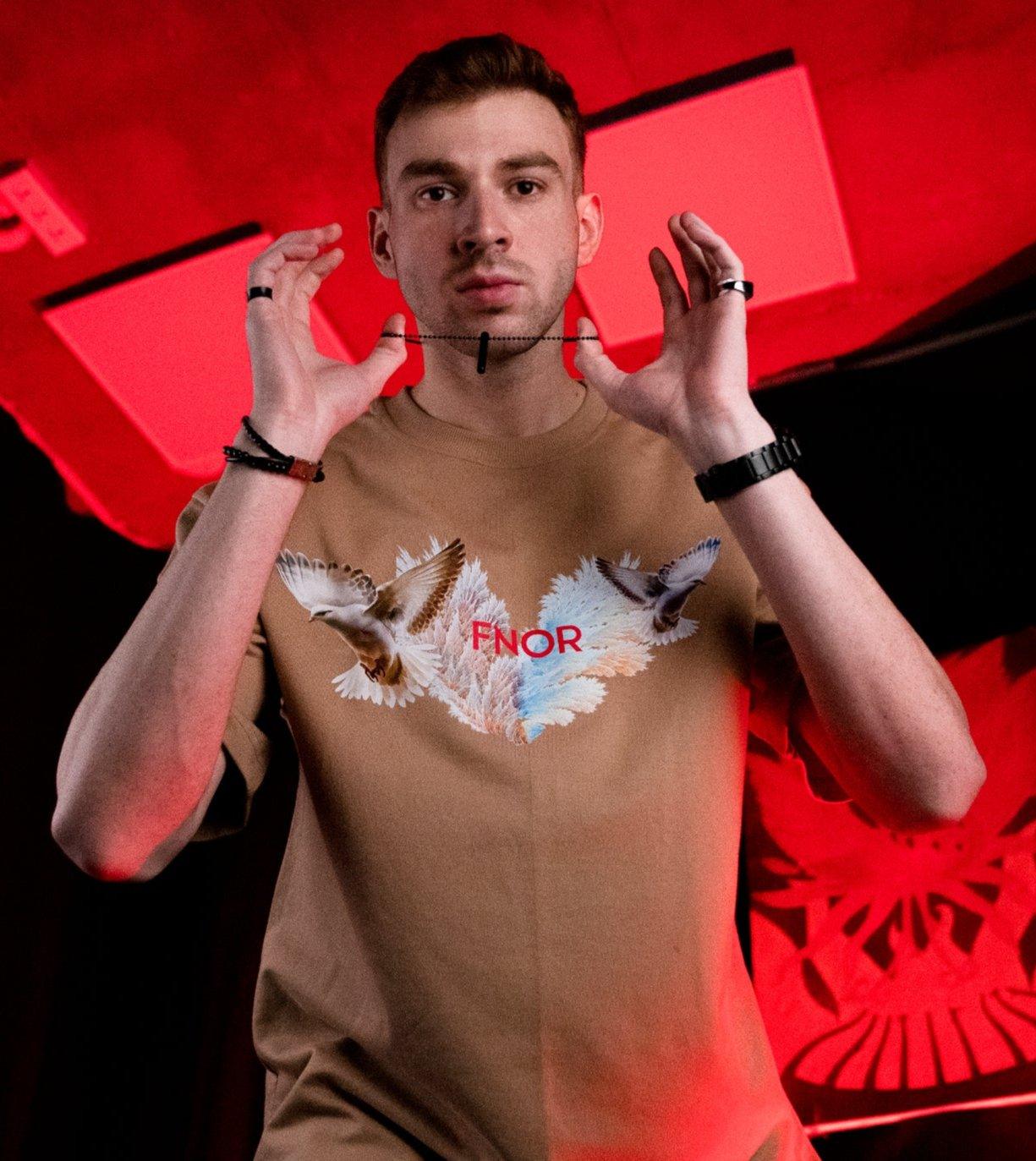  A man posing confidently in the FLY HIGH BEIGE T-SHIRT by FNOR, showcasing the front design with birds and the FNOR logo.
