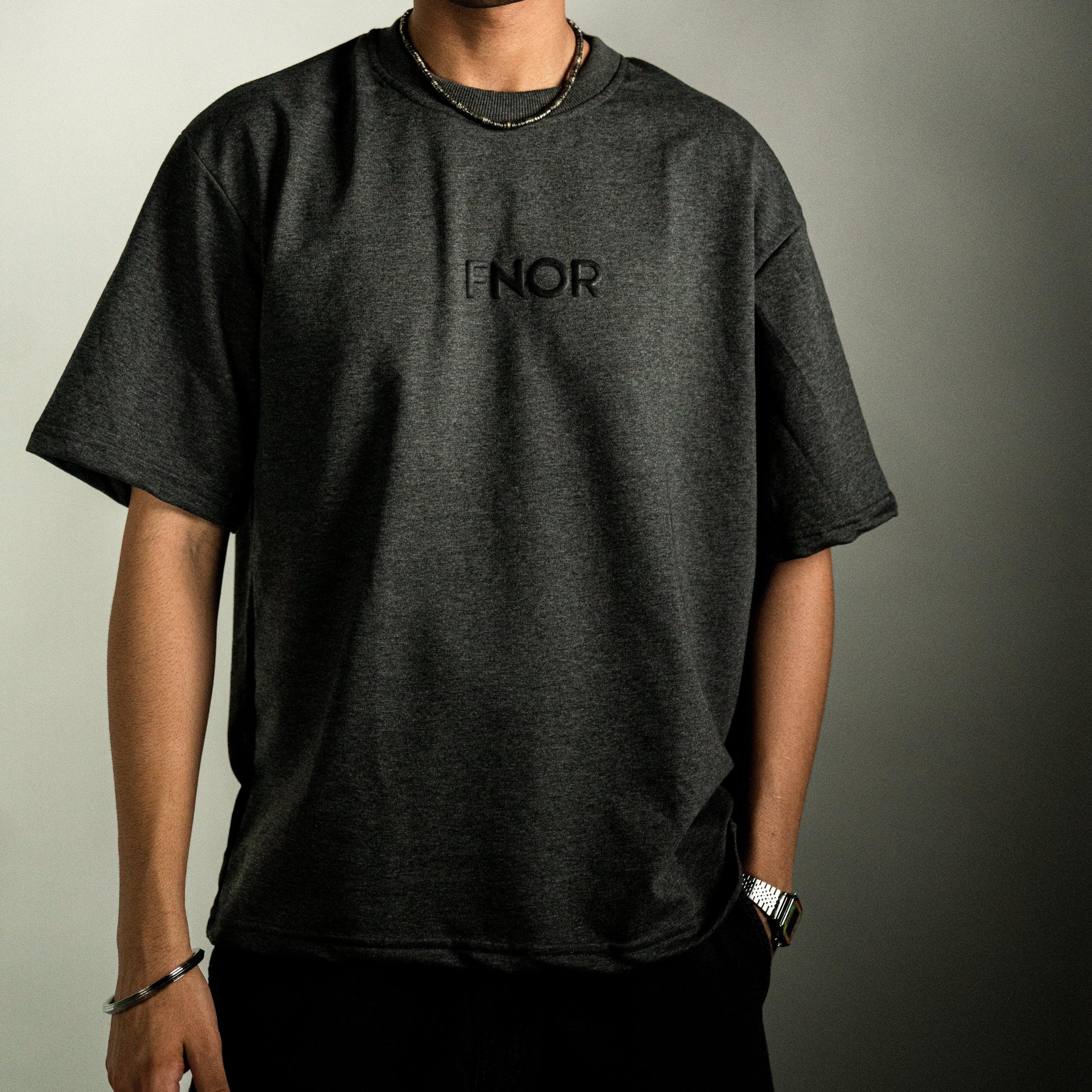 A person standing casually in the STONED ASH GREY T-SHIRT by FNOR, displaying the minimalist “FNOR” logo in black puff print on the front.